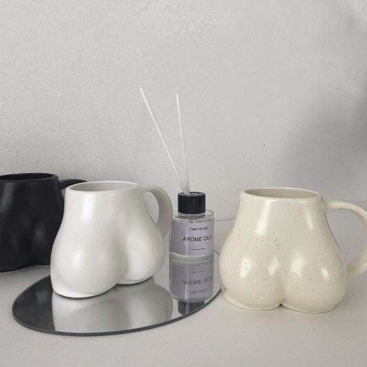 Two ceramic mugs and a diffuser on a reflective surface with a neutral background